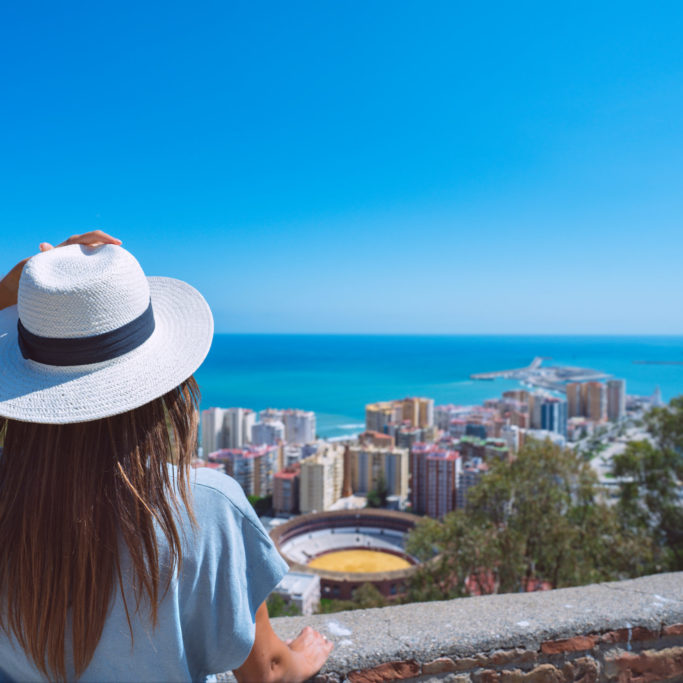 Rear view of young woman traveller with white hat standing in Gibralfaro Castle viewpoint and looking at Malaga cityscape. Summer holiday vacation in Spain. High quality photo